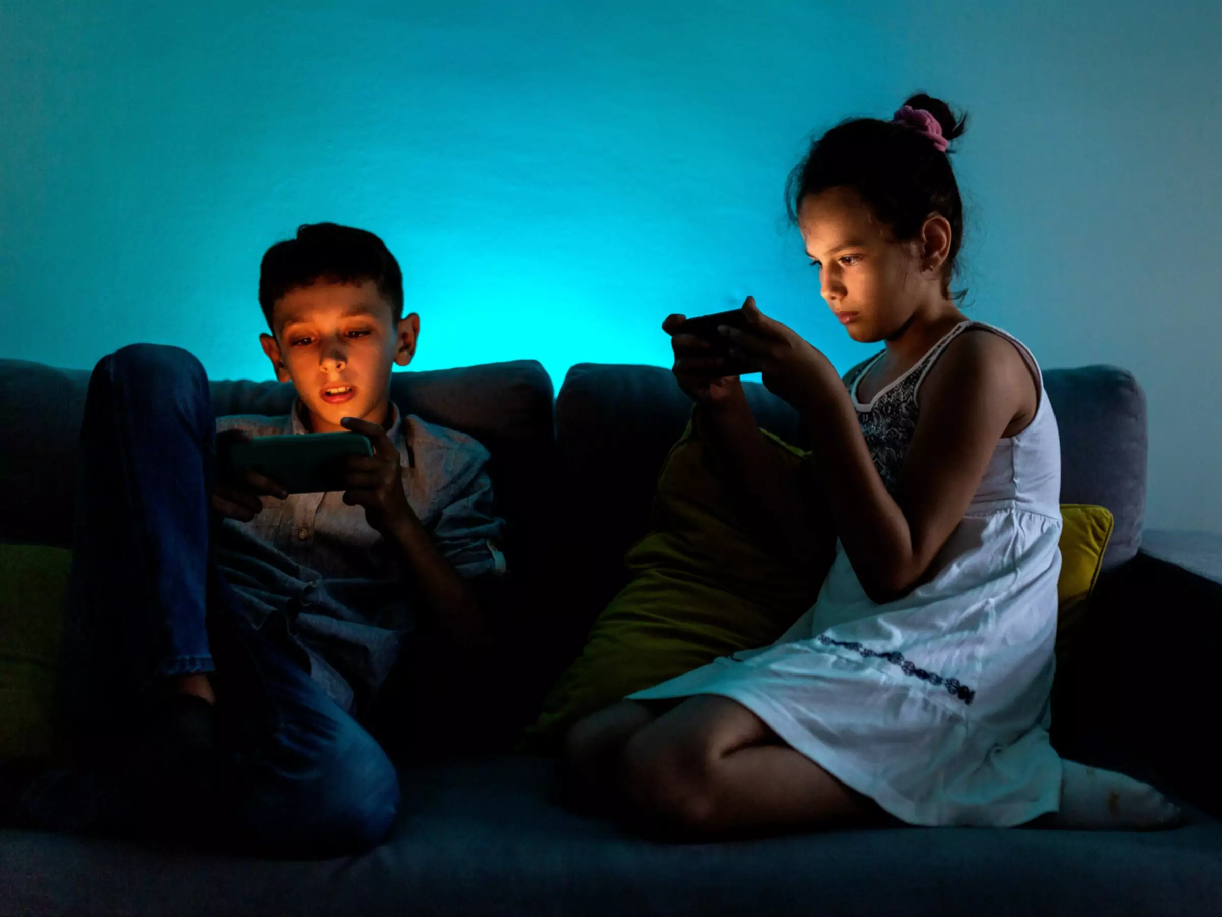Doi copii mici care stau pe canapea și se joacă jocuri pe smartphone-uri
